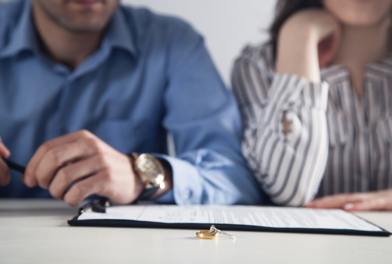 Image is of a married couple sitting at a table with divorce papaers in front of them, also in the foreground of the photo is their wedding rings laying on the table, concept of contested vs uncontested divorce