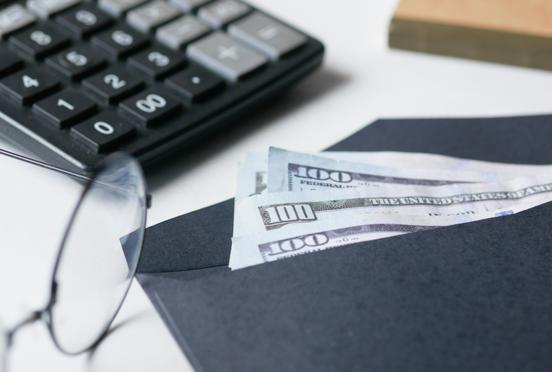 Image is of an envelope with multiple hundred dollar bills inside, on a table with a pair of glasses and a calculator, concept of how to calculate alimony in Tennessee