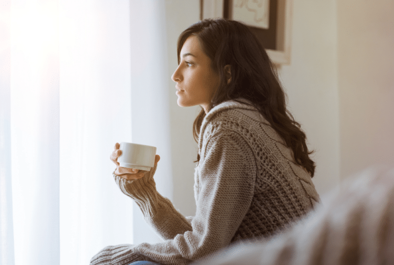 Image is of a young woman holding coffee cup and looking out the window while thinking, concept of signs it's time to hire a family lawyer in Franklin, TN