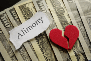Image is of 100 dollar bills fanned out on a table, a piece of paper on top that reads 'alimony' and a red felt heart that is broken in half, concept of alimony in a short-term marriage in Franklin, Tennessee