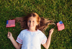 Image is of a little girl laying on green grass with little American flags in each hand, concept of 4th of July during a divorce in Franklin