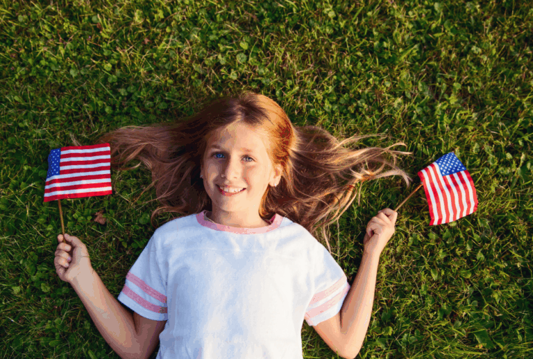 Image is of a little girl laying on green grass with little American flags in each hand, concept of 4th of July during a divorce in Franklin