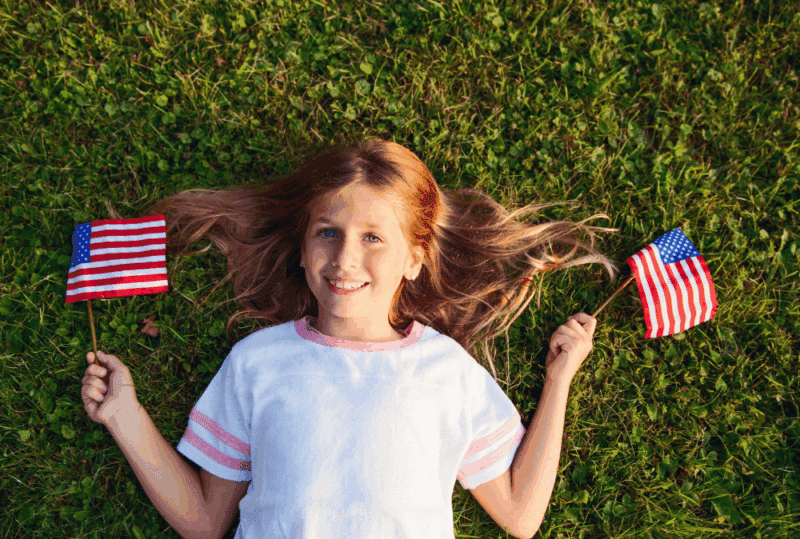 Image is of a little girl laying on green grass with little American flags in each hand, concept of 4th of July during a divorce in Franklin