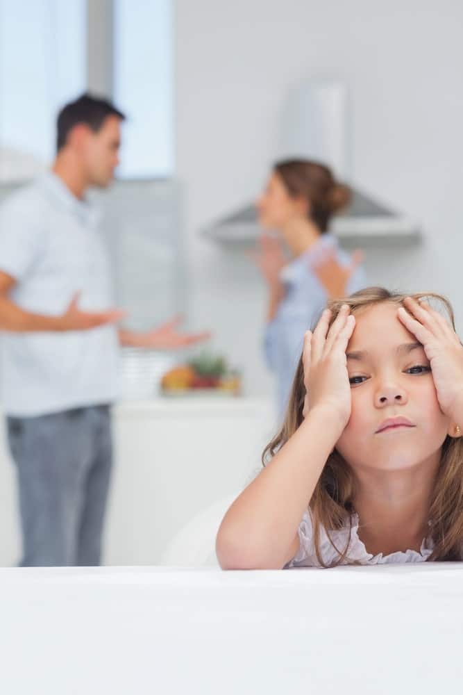 Image shows a frustrated child sitting with her head in hands while parents argue in the kitchen, symbolizing emotional impact of custody conflicts.