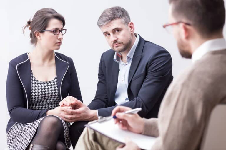 Image is of a man and woman holding hands while speaking with a professional taking notes on a clipboard, concept of divorce mediation