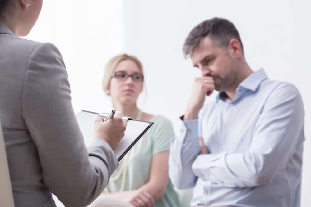 Image is of a legal professional writing on a clipboard while a man and woman look on thoughtfully, concept of divorce mediation