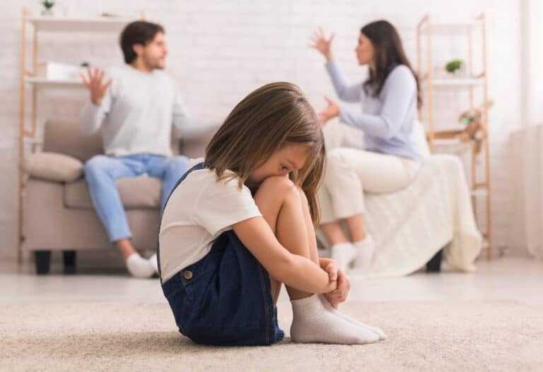 Image is of a child sitting alone while parents argue in the background, showing emotional impact of a school choice dispute on children