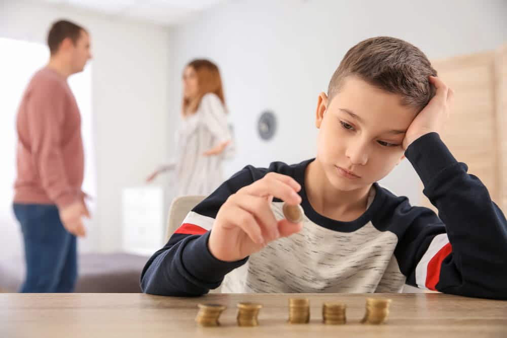 Image is of a child sitting alone while counting coins as parents argue in the background during a family dispute, concept of Franklin child support lawyer helping families manage financial and custody concerns.