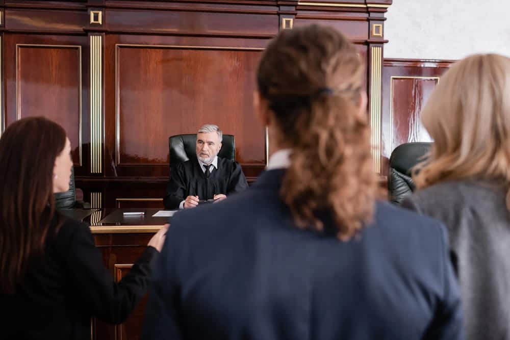 Image is of a judge addressing individuals inside a courtroom during a legal hearing, concept of court proceedings and legal disputes involving a custody order violation.