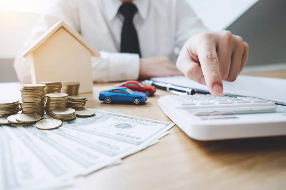 Image is of a person using a calculator beside money, a home model, and toy cars, concept of dividing credit card debt during divorce proceedings.