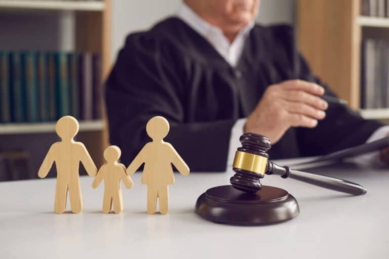 Image is of a judge seated behind a gavel with wooden figures representing parents and a child, concept of a custody hearing involving child custody and family court decisions.