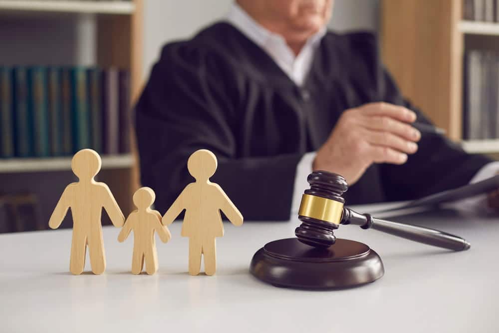 Image is of a judge seated behind a gavel with wooden figures representing parents and a child, concept of a custody hearing involving child custody and family court decisions.
